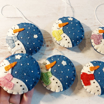 A set of six round shaped felt ornaments with smiling snowmen in blue and white colors, each with a different color accent on the top, displayed on a white surface.