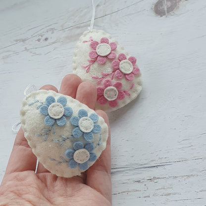 Two felt egg ornaments with floral patterns, one in pink and one in blue, held in a hand against a white background.