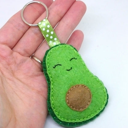 Avocado-shaped keychain with a smiling face held in a hand against a white background